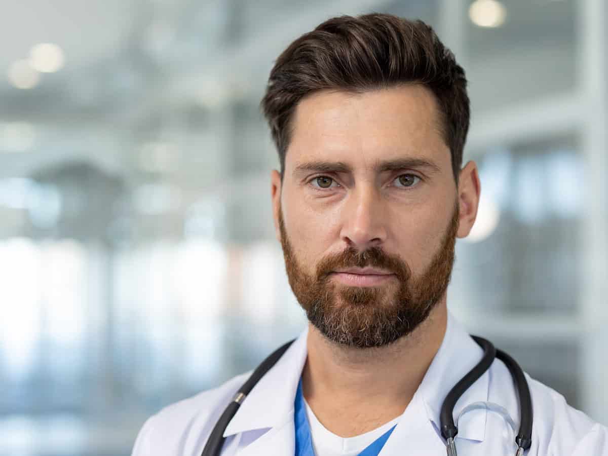close up portrait of serious experienced doctor, man thoughtfully looking at camera in white medical coat, working inside modern clinic office