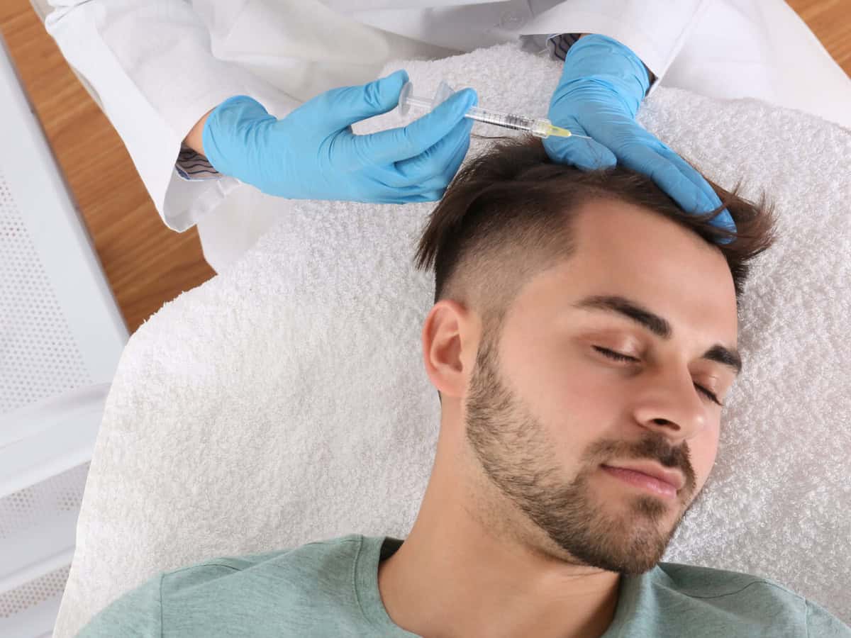 Thailand or Turkey? Compare Hair Transplant Surgeon Quality young man with hair loss problem receiving injection in salon, top view